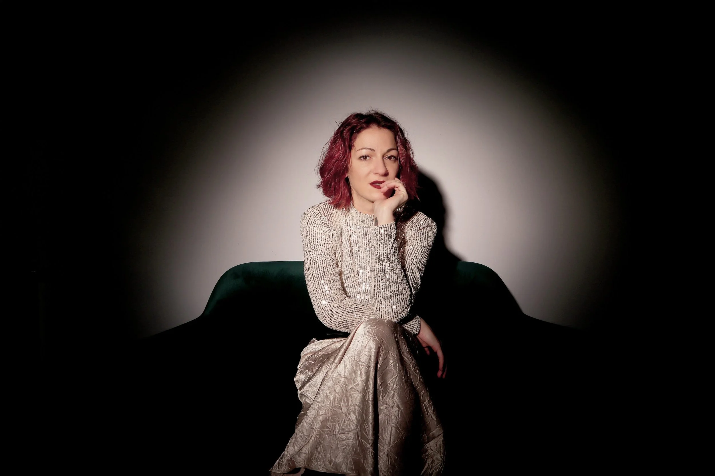Eine Frau mit roten Locken und dunkler Lippenfarbe sitzt auf einem dunkelgrünen Sofa vor einem weißen Hintergrund. Sie trägt einen silberfarbenen, glänzenden Pullover und eine ebenfalls glänzende lange Hose. Sie ruht ihren Kopf auf ihrer rechten Hand und schaut nach vorne.