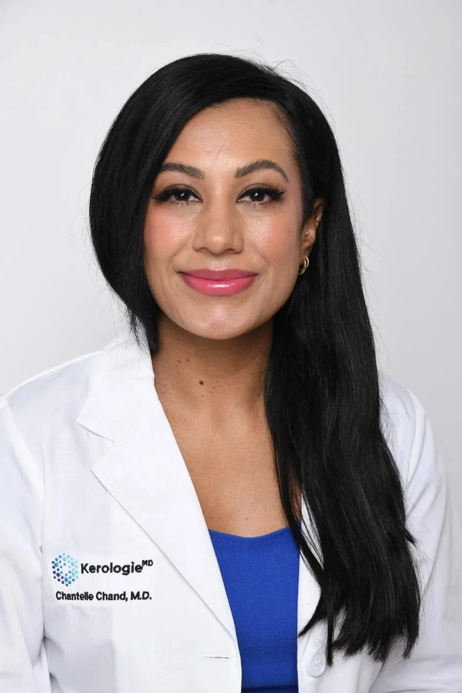 A woman with long black hair wearing a white coat and a blue top, smiling.
