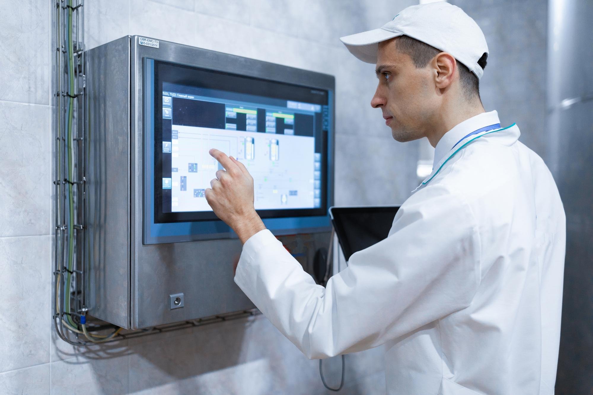 A scientist in a lab coat and cap working on a touchscreen panel in a laboratory.