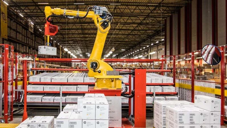 Industrial robotic arm within a warehouse, handling boxes of product surrounded by safety barriers arranging onto pallets.