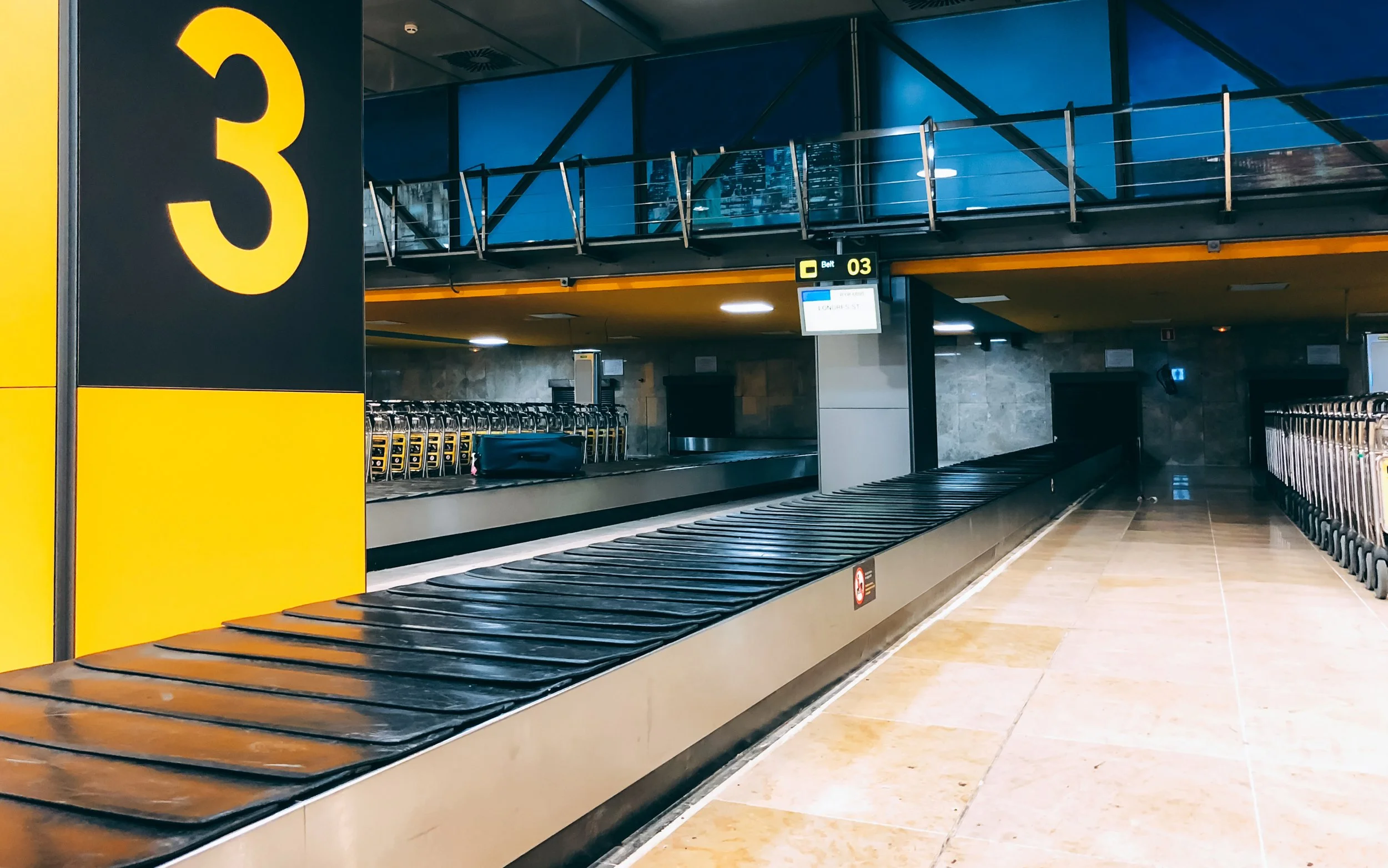 Empty baggage claim area at an airport with a large yellow and black number 3 sign, luggage conveyor belt, and carts in the background.