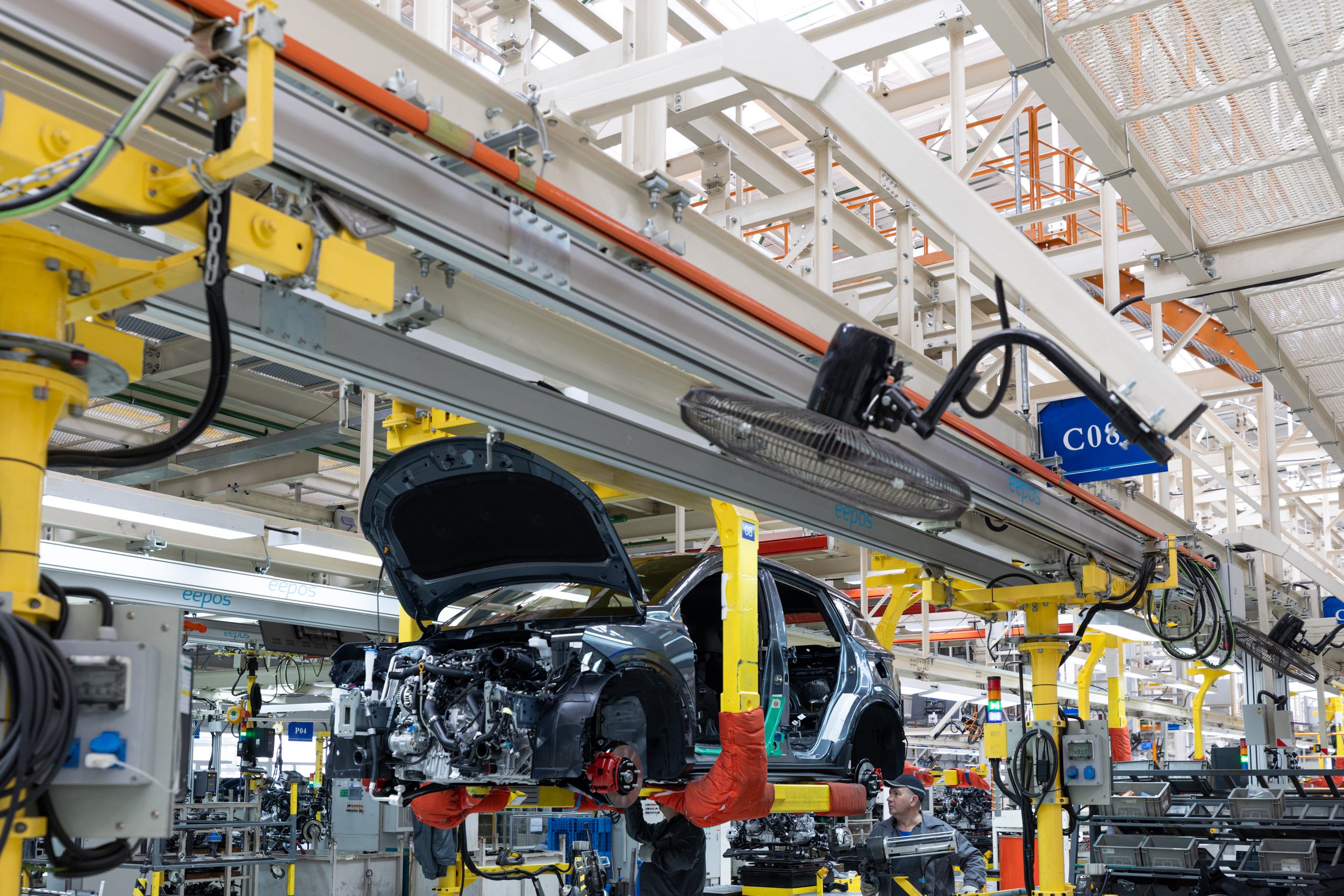 Automobile assembly line with a partially assembled car being worked on by technicians in a factory. The car's engine and inner parts are visible, and the surrounding machinery and frameworks are painted yellow, white, and orange.