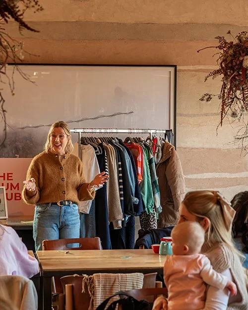 A woman standing in front of a clothing rack filled with jackets and shirts, speaking to a group of people seated at a table, with a baby in a woman’s lap.
