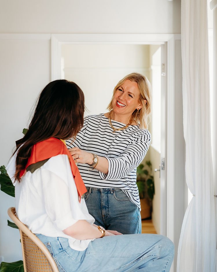 A woman with blonde hair smiling and talking to another woman with dark hair, who is sitting. They are inside a room with white curtains and a mirror in the background.