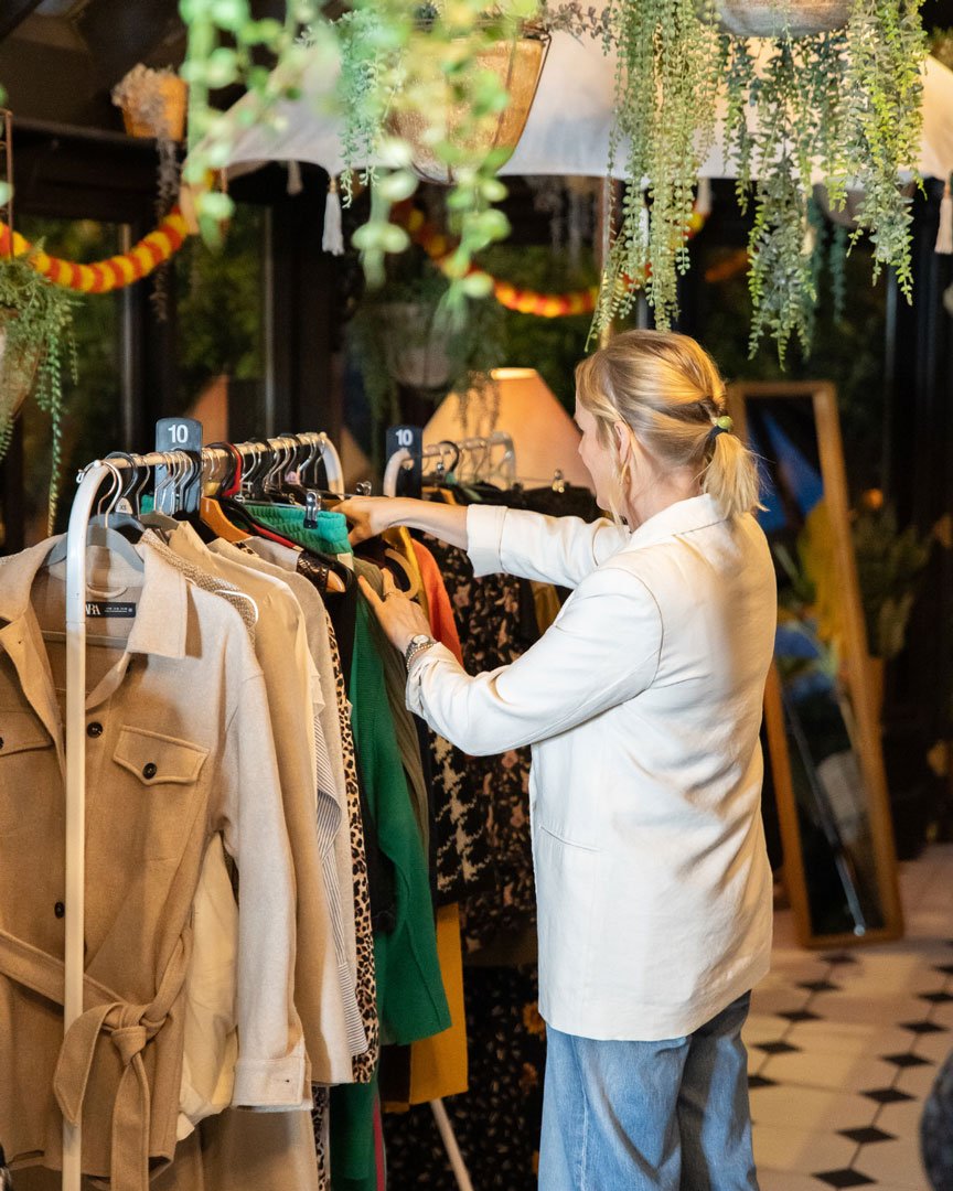 A woman shopping for clothes at an indoor boutique store, browsing a rack with various colorful garments including beige, green, leopard print, and black floral patterns, surrounded by green hanging plants and warm lighting.
