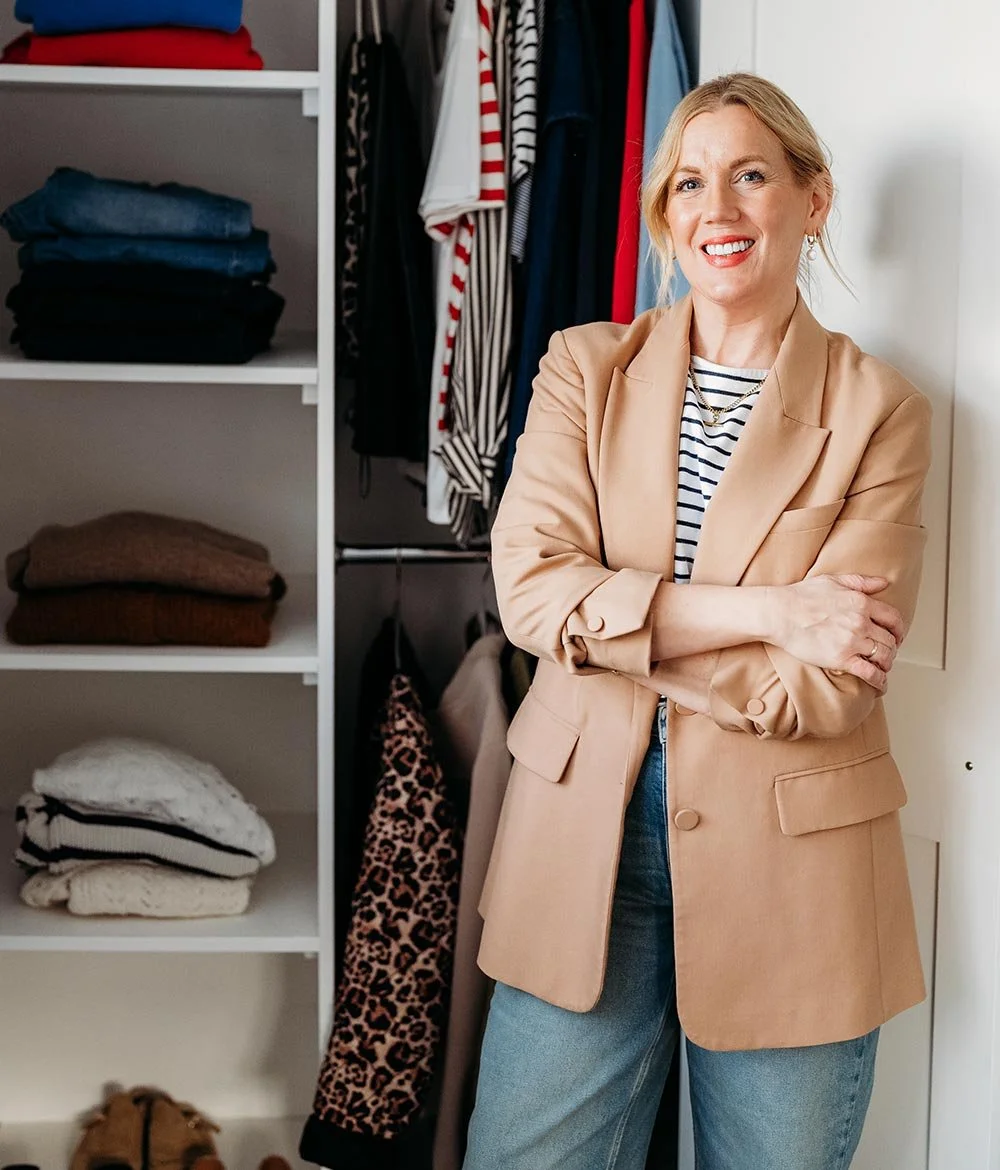 A woman smiling with arms crossed standing in front of a walk-in closet with folded clothes and hanging garments.