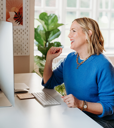 A woman smiling and celebrating while looking at her computer screen in a bright, cozy home office.