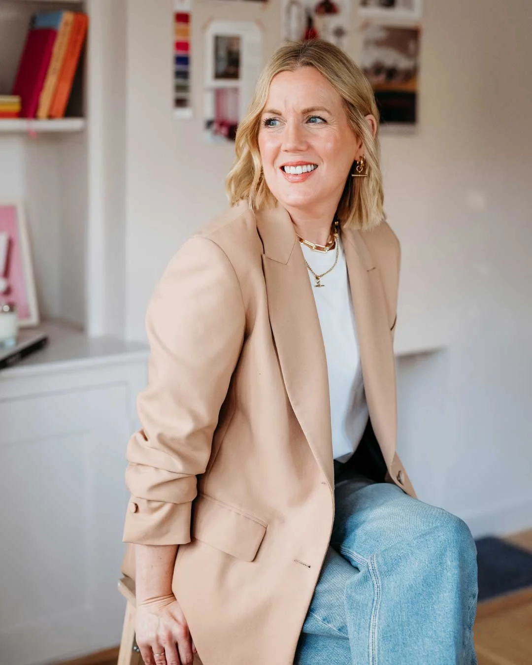 A woman with blonde hair, wearing a beige blazer, white shirt, and blue jeans, sitting relaxed with a smile in a room with bookshelves in the background.