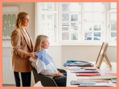 A woman standing behind a seated woman in a dentist's chair, with a blue dental bib, in a well-lit room with large windows and a desk with papers and a mirror.