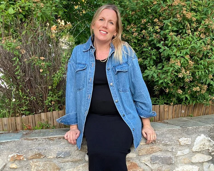 Blonde pregnant woman sits on a wall infrint of a green bush wearing a denim short and black dress