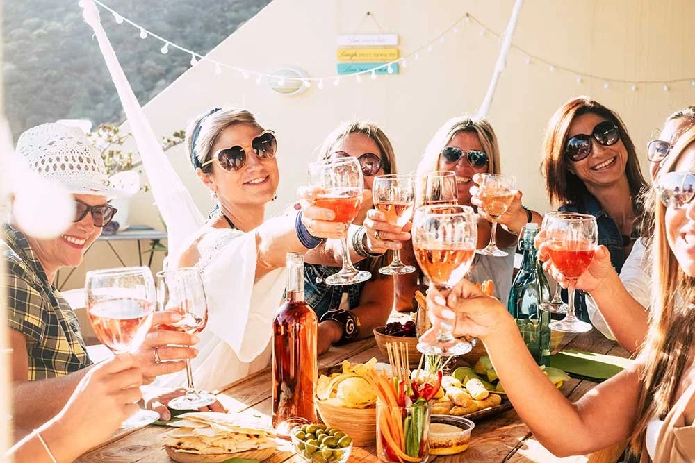 Group of women celebrating outdoors at a picnic table with drinks and snacks under a canopy with string lights.