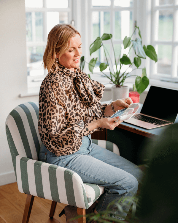 A woman with blonde hair sits on a striped chair in a bright room, holding a tablet, with a laptop on the table beside her and a large potted plant nearby.