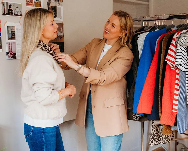 Two women stand infront of a rail of clothes. One woman adjusts the clothing of the second woman