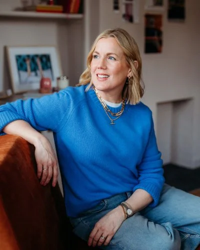 Woman with blonde hair wearing a blue sweater and jeans sitting on a sofa in a room with bookshelves and framed photos