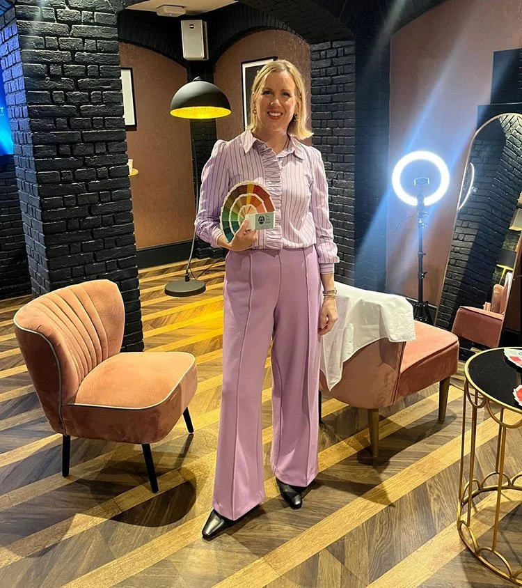 A woman in pink pants and a striped blouse holding a color palette in a stylish indoor setting with black brick walls, wooden flooring, and modern furniture.