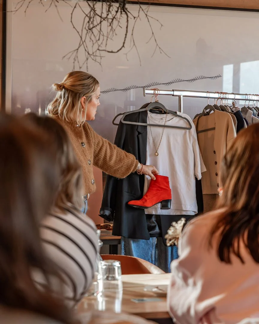 A woman is showing a red ankle boot to a group of people seated at a table, with clothing and accessories hanging on a rack behind her.