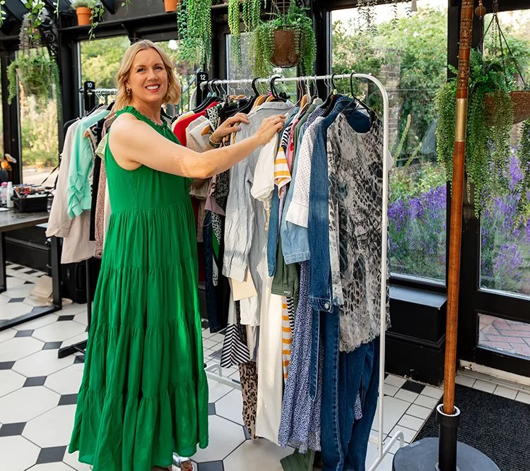 A woman with blonde hair wearing a green dress is shopping for clothes at a boutique, browsing a clothing rack filled with various garments.