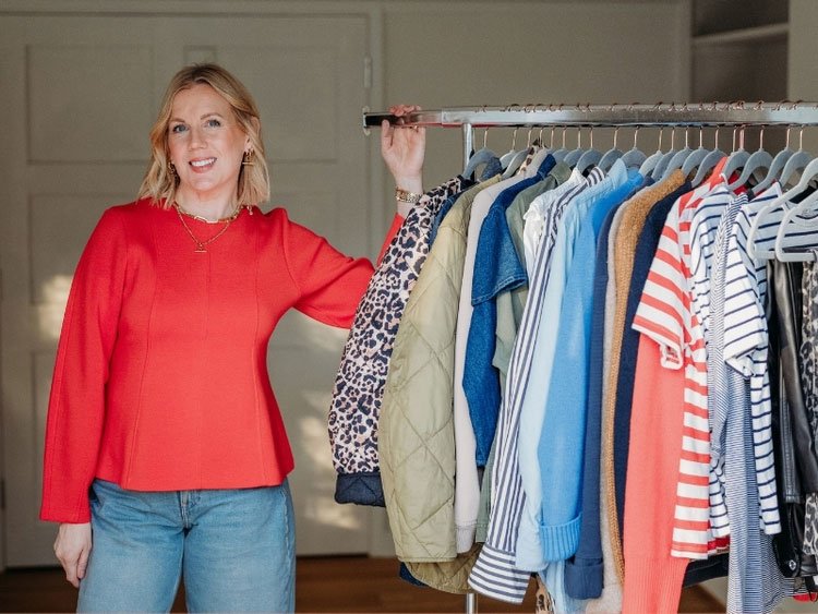 Woman smiling in red sweater standing next to a clothing rack with various colorful shirts and jackets.