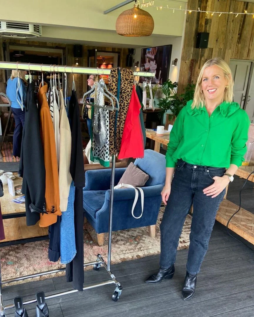 A smiling blonde woman wearing a bright green blouse and black jeans standing next to a clothing rack with various garments in a boutique or fashion store.