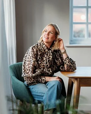 A woman with blonde hair sitting at a wooden table by a window in a room filled with natural light, wearing a leopard print blouse and blue jeans.