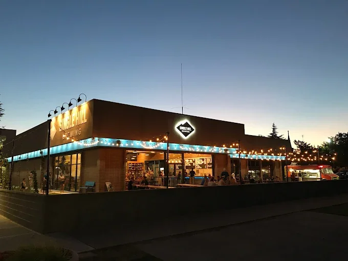 Exterior of a Black Hills Brewing restaurant during evening, with large windows, string lights, and a lit sign, showing customers inside and a menu board outside.