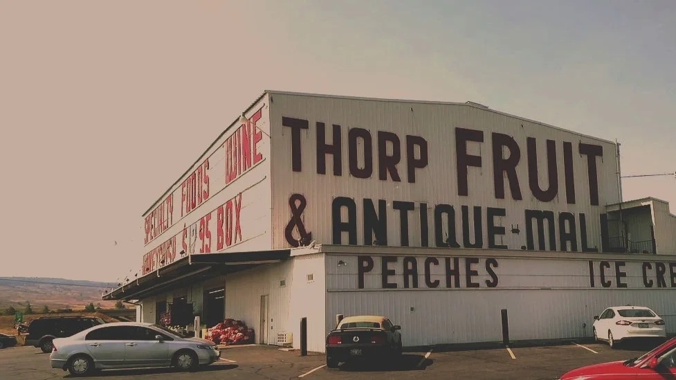 A large building with signs indicating it sells Thorp fruit and antique mall, peaches, and ice cream. There are several parked cars in front of the building, with a clear sky in the background.