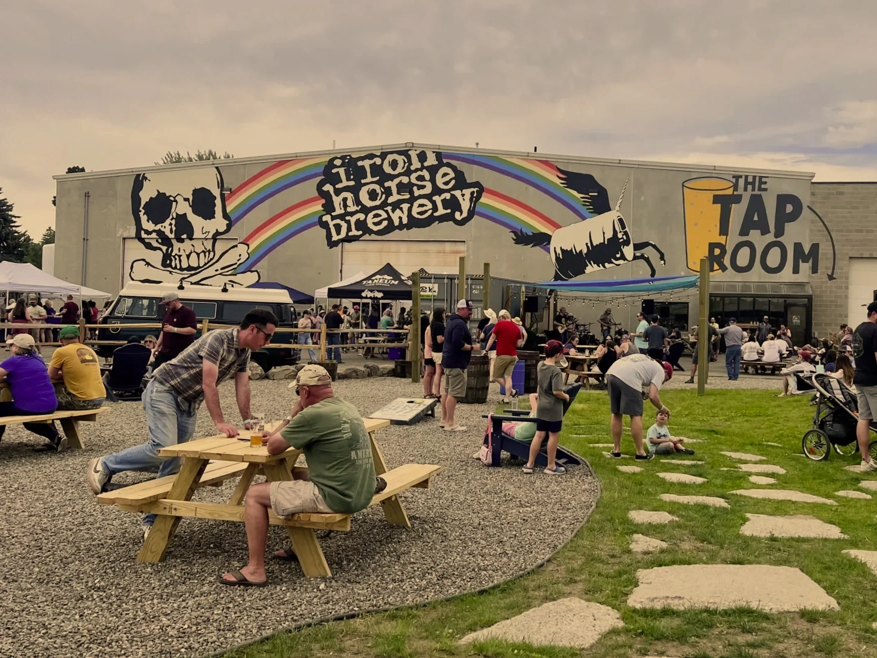 People gather outside Highland Horse Brewery's taproom with outdoor seating, wooden picnic tables, and a stage, under a mural of a skull with rainbow, featuring a skeleton horse and a giant beer glass.