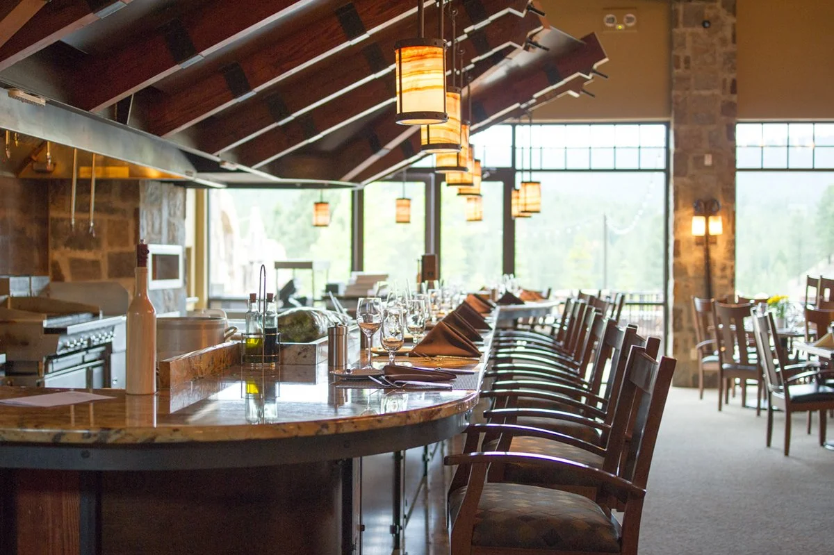 Interior view of a restaurant bar with empty chairs, glasses, and place settings, illuminated by hanging pendant lights and large windows with a view outside. Suncadia