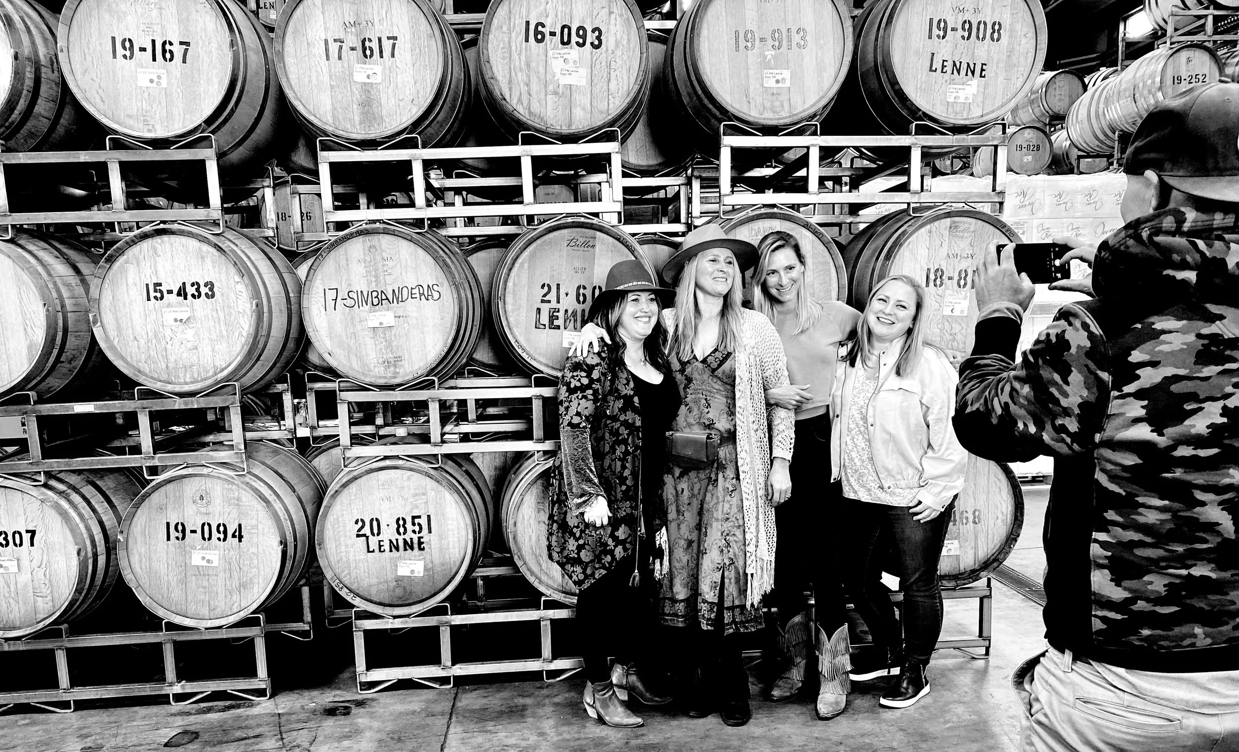 Four women standing in front of stacked wine barrels, smiling for a photo being taken by a man in a camouflage jacket.