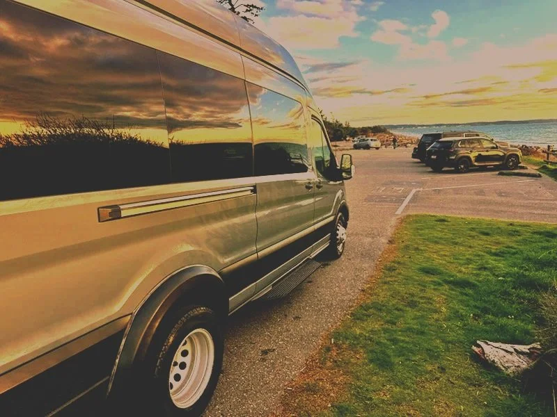 A parked beige van near a grassy area with a view of the ocean and several other vehicles at a coastal parking lot during sunset.