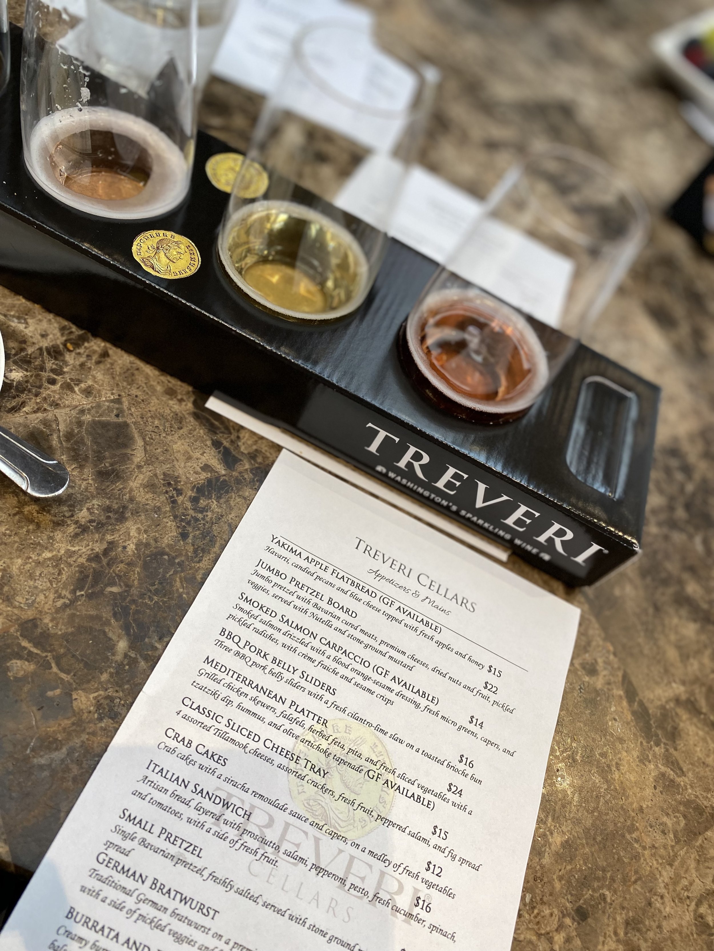 A tasting flight of four small glasses of beer in a black tray labeled 'Treveri' on a marble table, with a printed menu of appetizers and mains underneath.