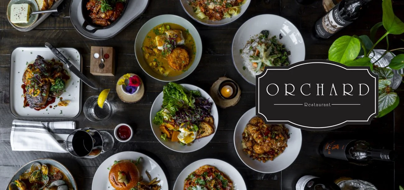 A top-down view of a table filled with various plates of food, drinks, and small decorative items, with a sign that reads "Orchard Restaurant" on the right side of the image.