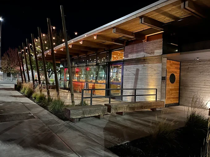 Nighttime view of a modern building with glass walls, wooden accents, and outdoor seating. Decorative vertical wooden poles and benches are outside along a sidewalk with planter boxes.