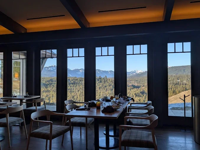 Interior of a dining area with a long table and chairs, large windows showing a mountain landscape with snow-capped peaks and dense forest, during daytime. Suncadia