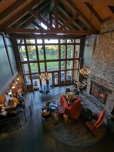 Interior view of a spacious living room with high vaulted wooden ceilings, large windows, a stone fireplace, and cozy seating with a red sofa and armchairs. Suncadia