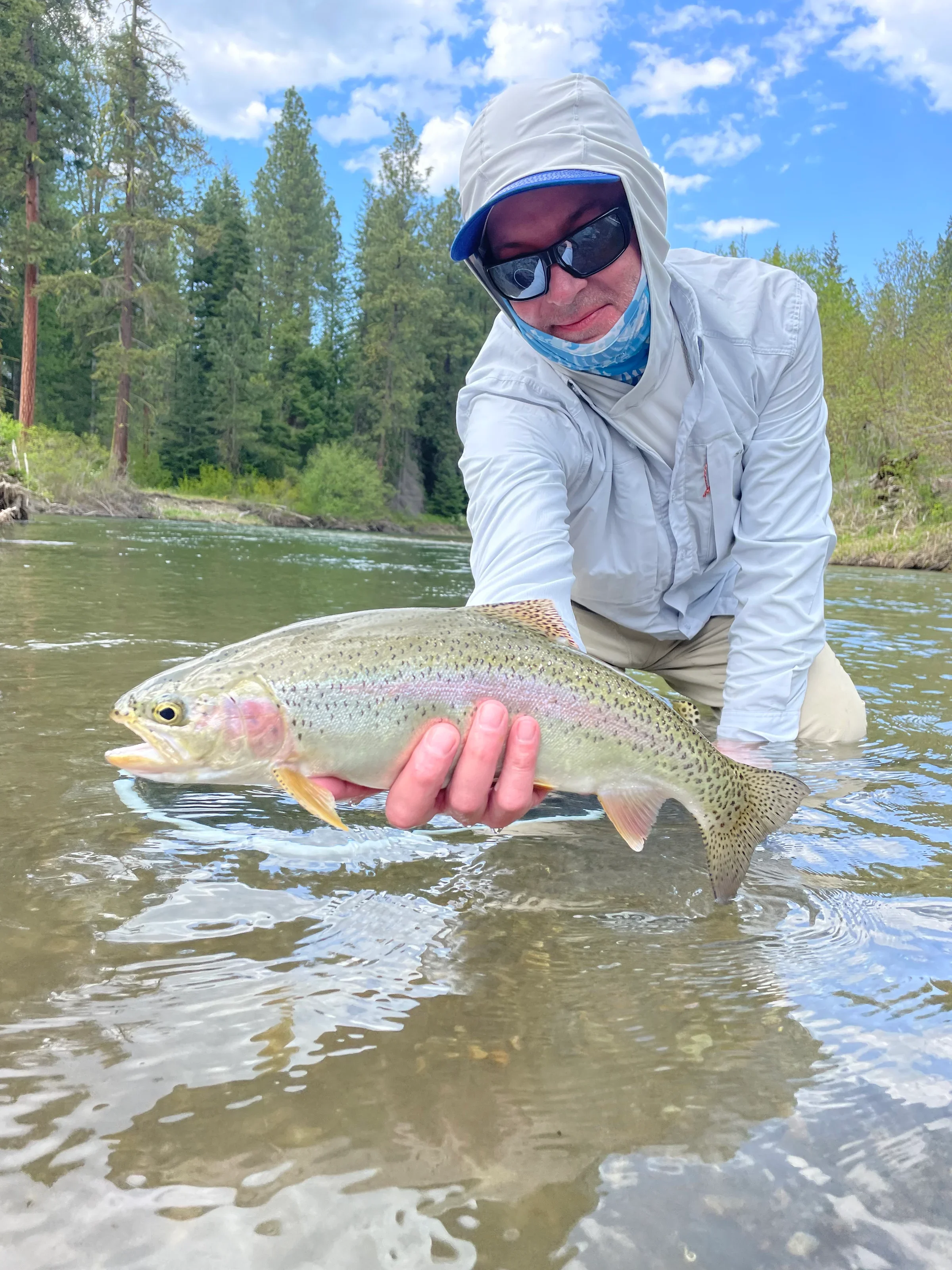 Man wearing sunglasses, a hoodie, and a face mask, holding a large rainbow trout in shallow river water surrounded by trees and a partly cloudy sky. Guided Fly-Fishing Charters. Yakima River. Cle Elum