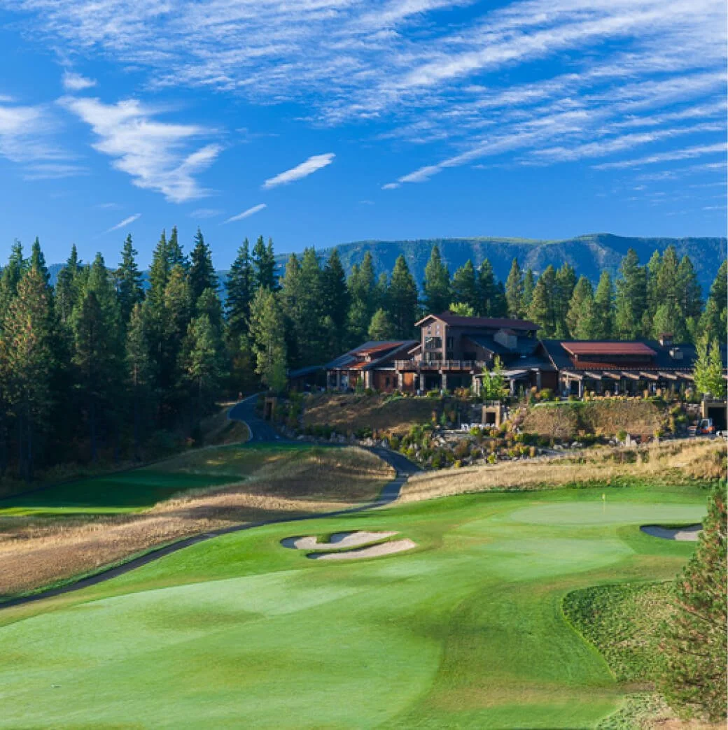 A golf course with a putting green in the foreground, a large house amid trees on a hillside, and mountains with a blue sky and scattered clouds in the background.