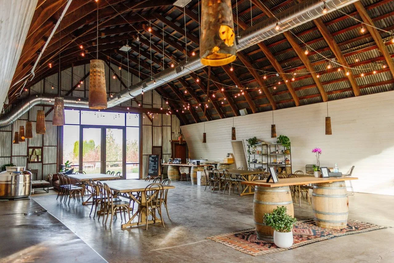 Interior of a modern rustic event space or restaurant with high arched wooden ceiling, string lights, large window, wooden tables, chairs, and decorative elements like plants and flowers. Gilbert winery. Winery tour