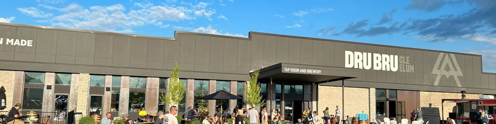 People gathering outside a modern building with a sign that reads 'DRU DRU' and 'TAP ROOM AND BREWERY' under a blue sky with clouds.