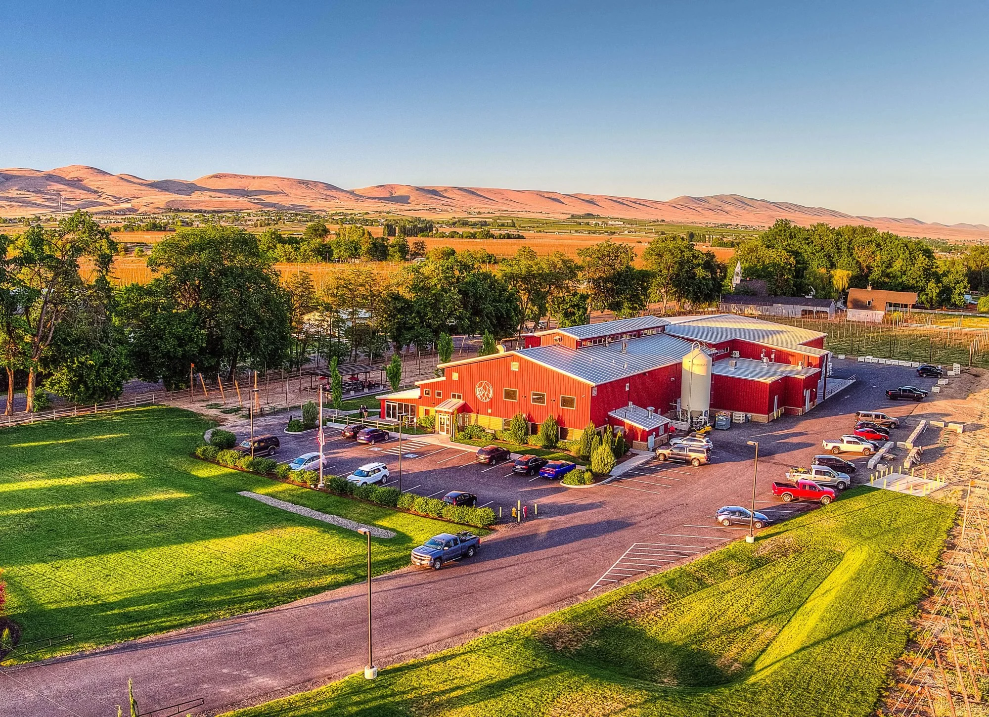 Aerial view of a large red barn with surrounding parking lot, green lawn, trees, and distant rolling hills under a clear blue sky. Bale Breaker yakima. Brewery tour. Bachelor party