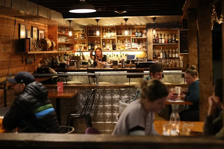 People sitting and chatting at tables in a cozy bar or café with a bartender behind the counter, shelves stocked with bottles of alcohol, and warm lighting.