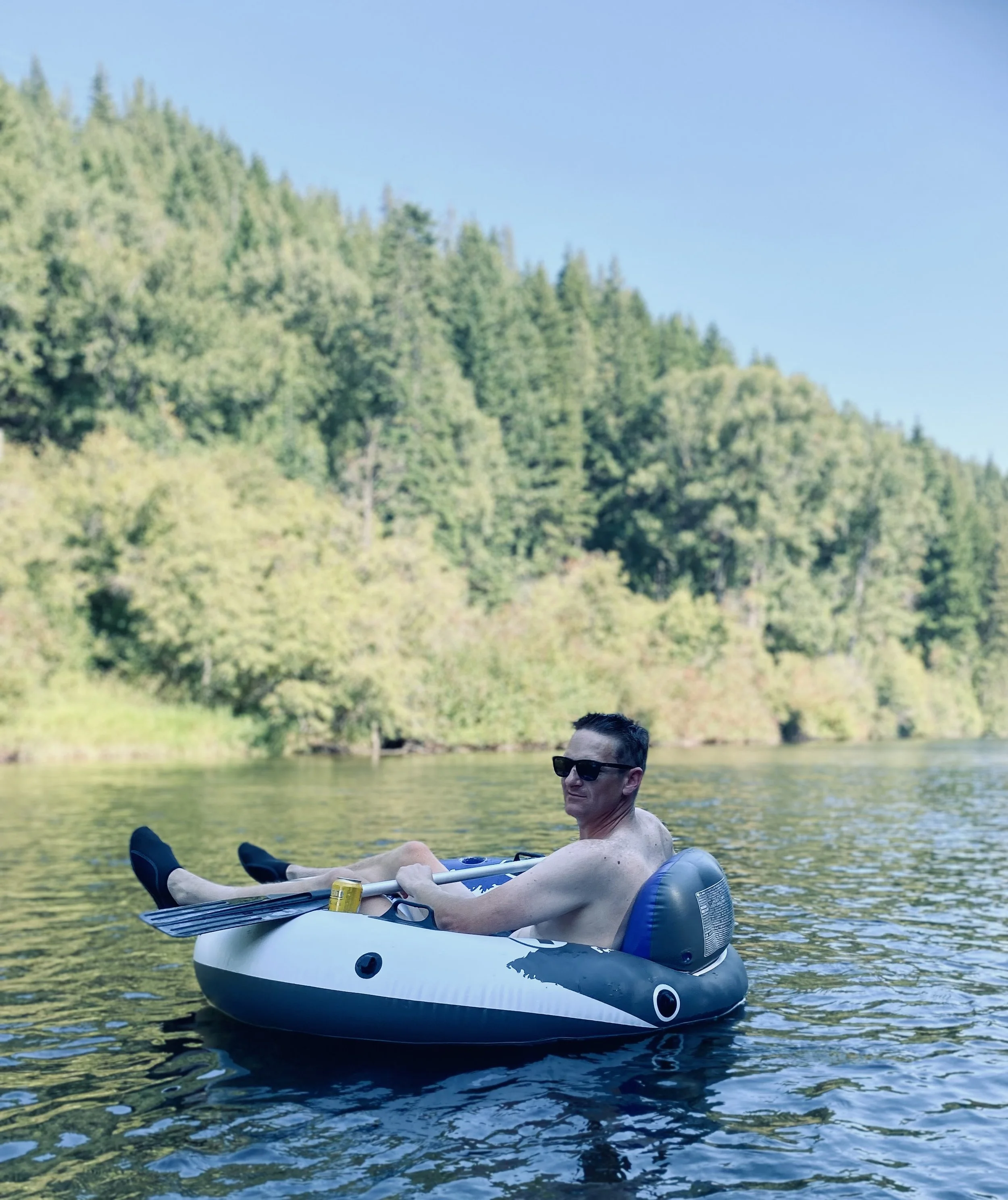 A man wearing sunglasses relaxing in an inflatable kayak on a lake surrounded by trees and mountains. river float cle elum. Roslyn. Suncadia