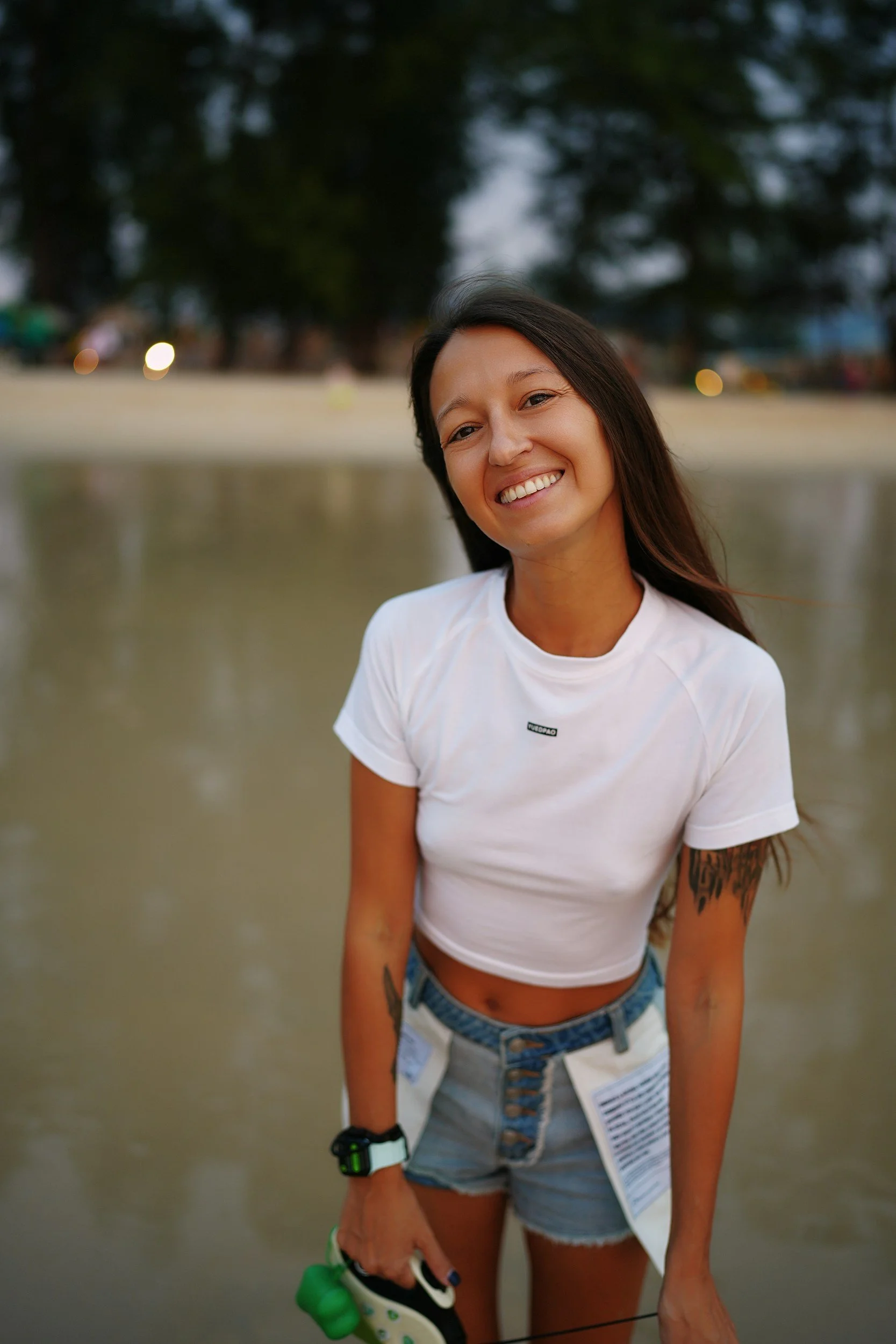 A woman smiling, standing near a body of water, wearing a white t-shirt, denim shorts, and a watch, with trees and a park in the background.