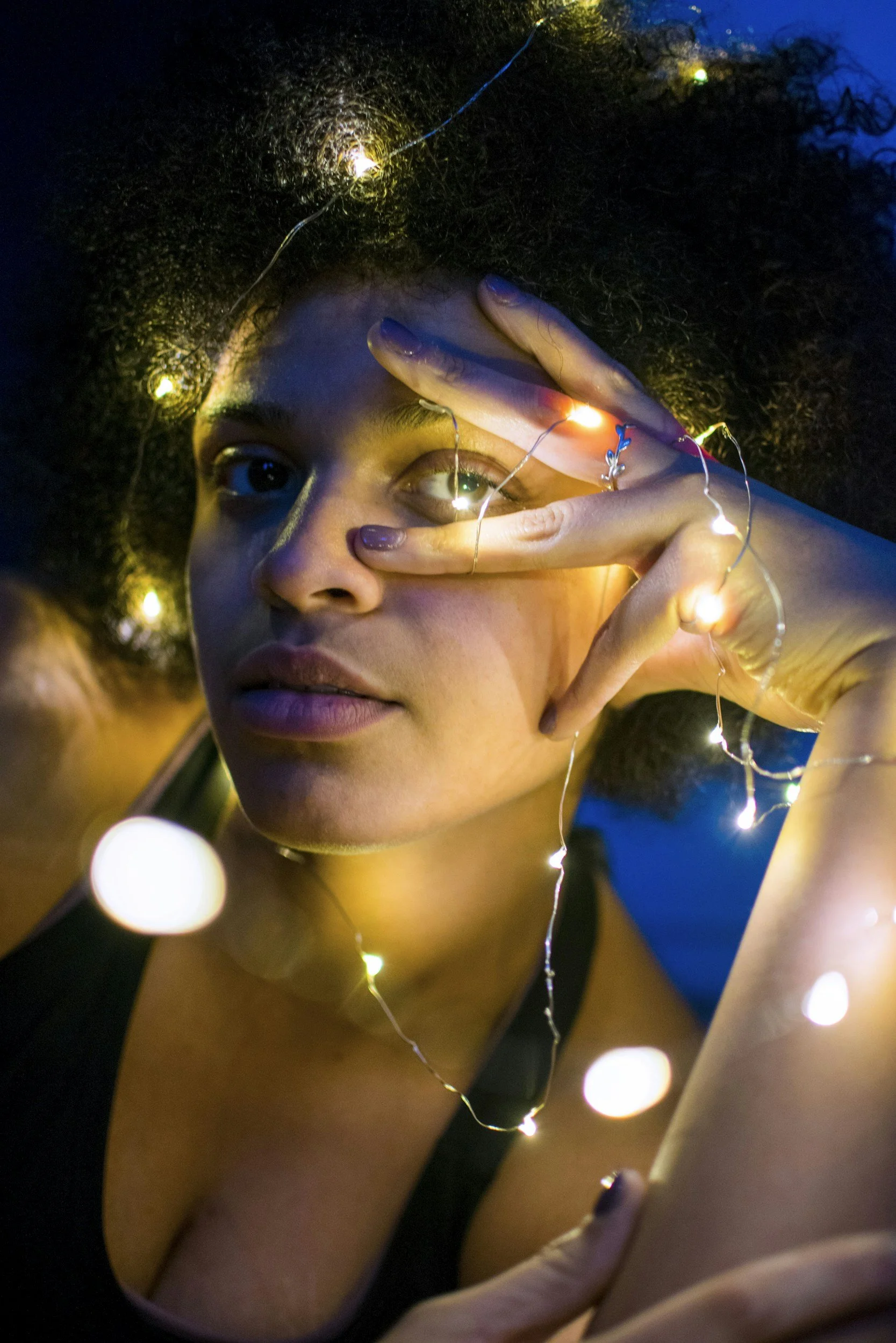 Close-up of a young woman with curly hair, holding her fingers in front of her face with fairy lights wrapped around her fingers and hair, creating a glow effect in a dark setting.