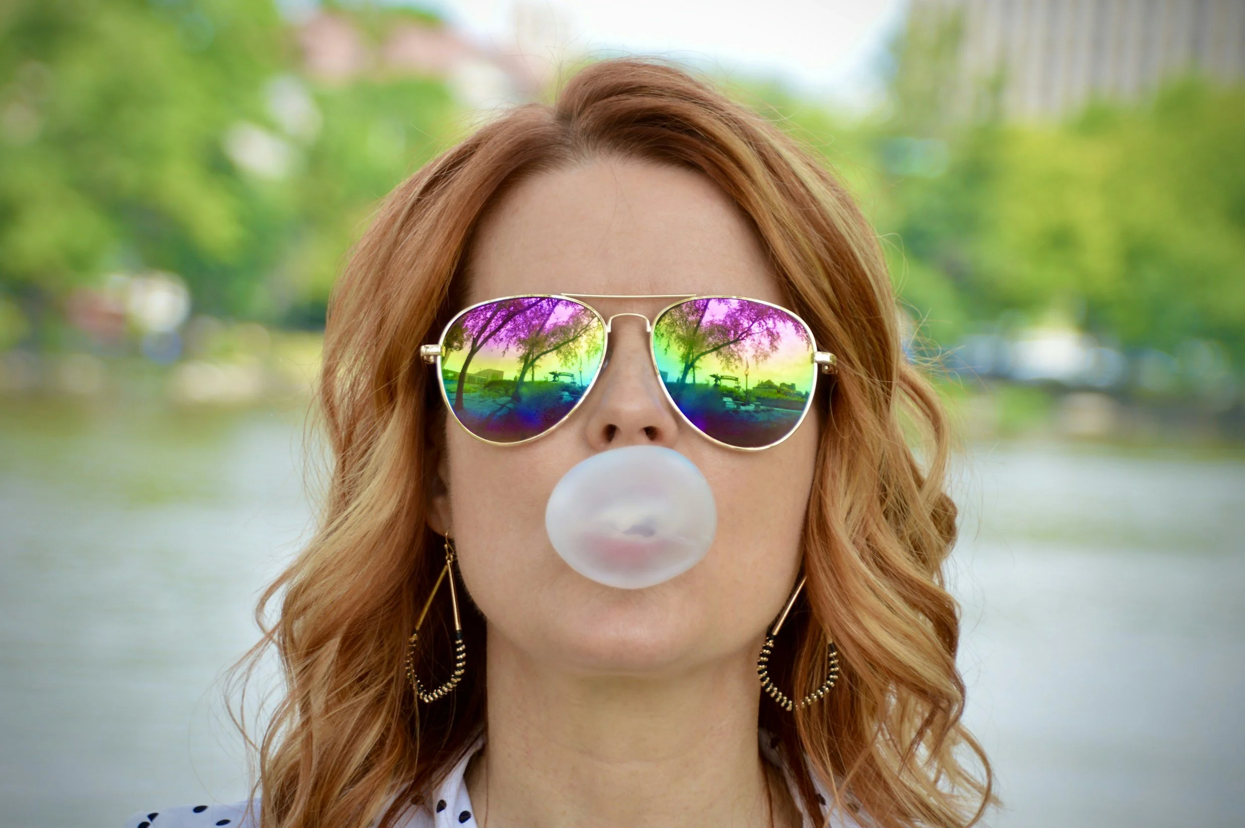 Woman with red hair wearing reflective sunglasses and blowing bubble gum, standing outdoors near a body of water with trees and buildings in the background.