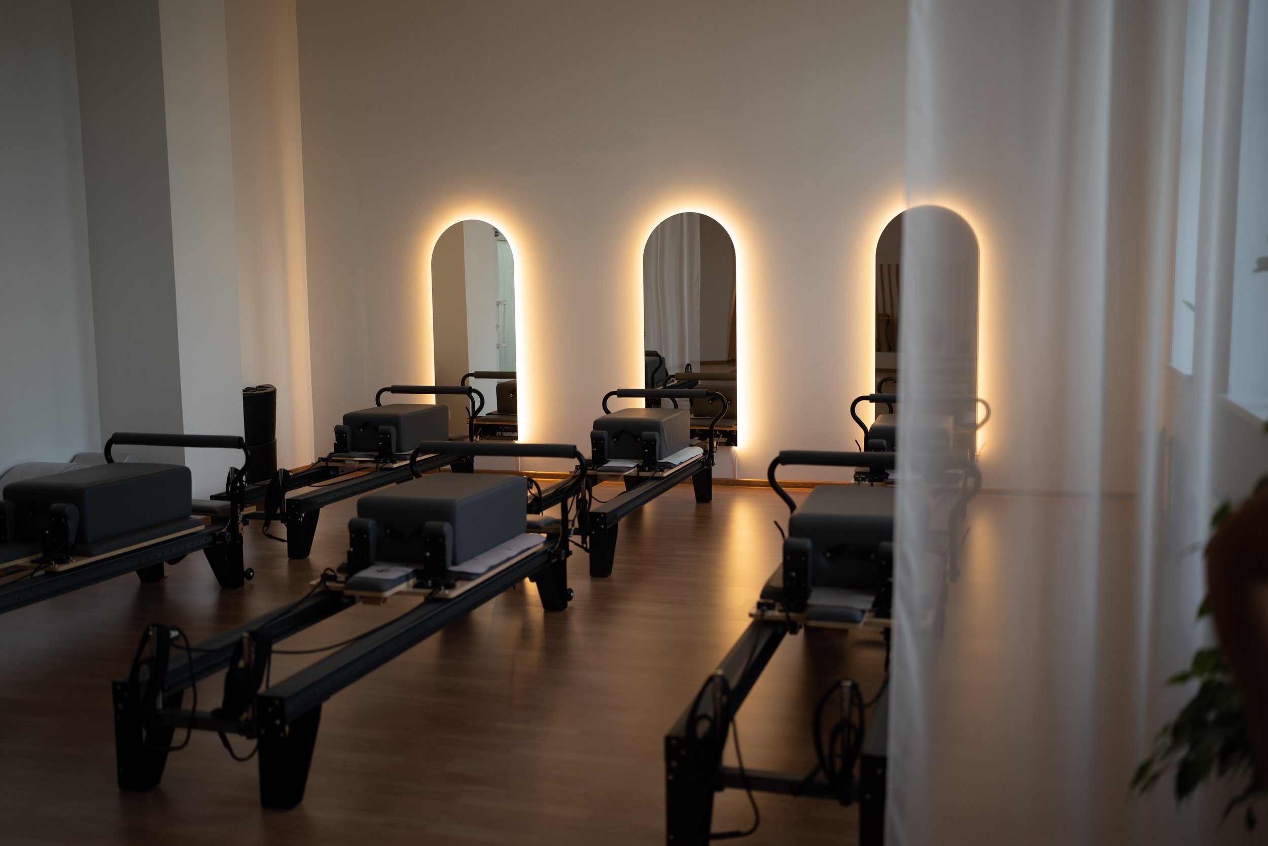 A Redormer Pilates studio with reformer Pilates machines arranged in rows, illuminated by soft backlighting along the wall with three illuminated arch-shaped mirrors.