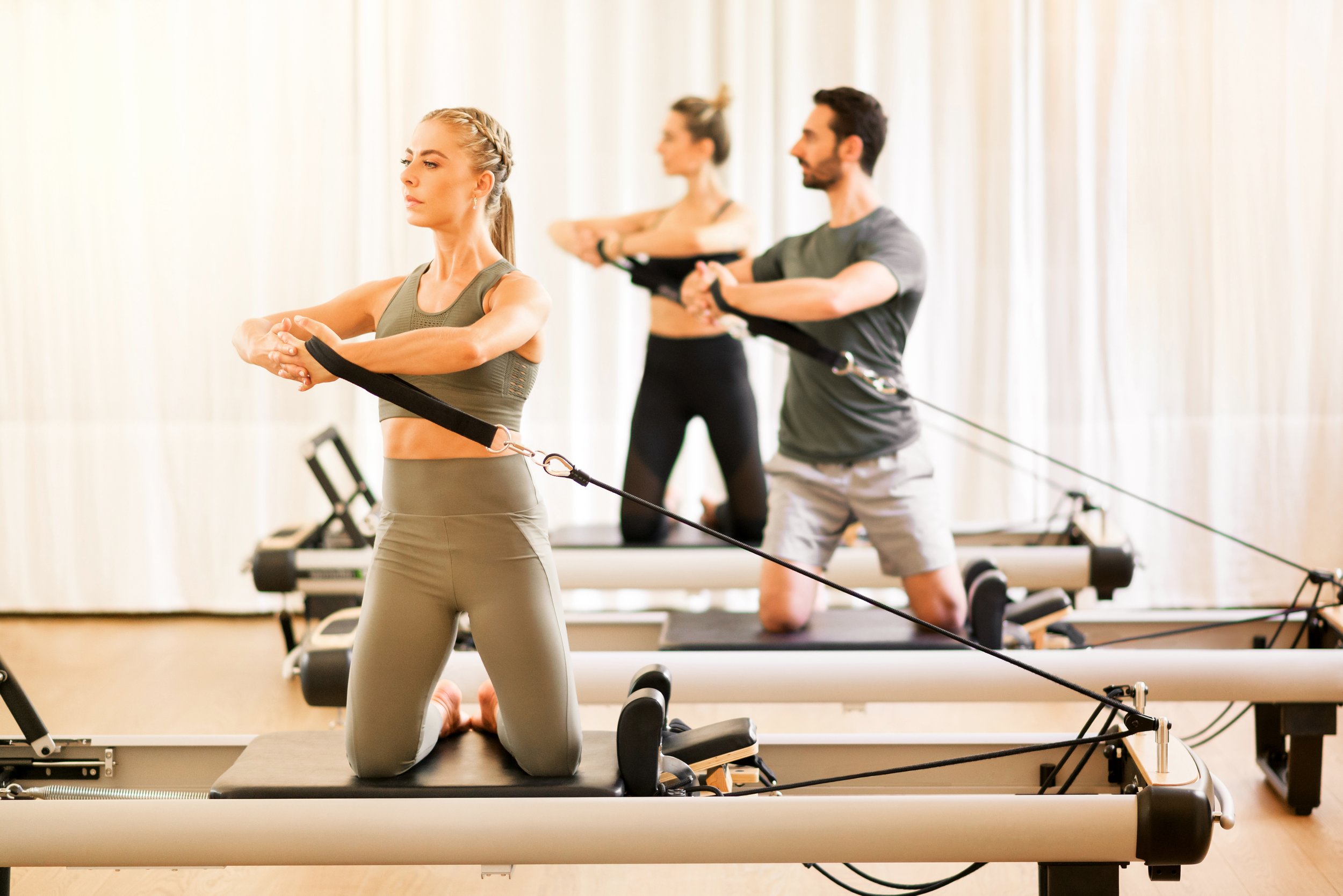 Three people practicing Pilates on reformer machines in a studio. The woman in front kneels on a machine pad, wearing sportswear, while the two people behind her, a woman and a man, are also exercising on reformers.