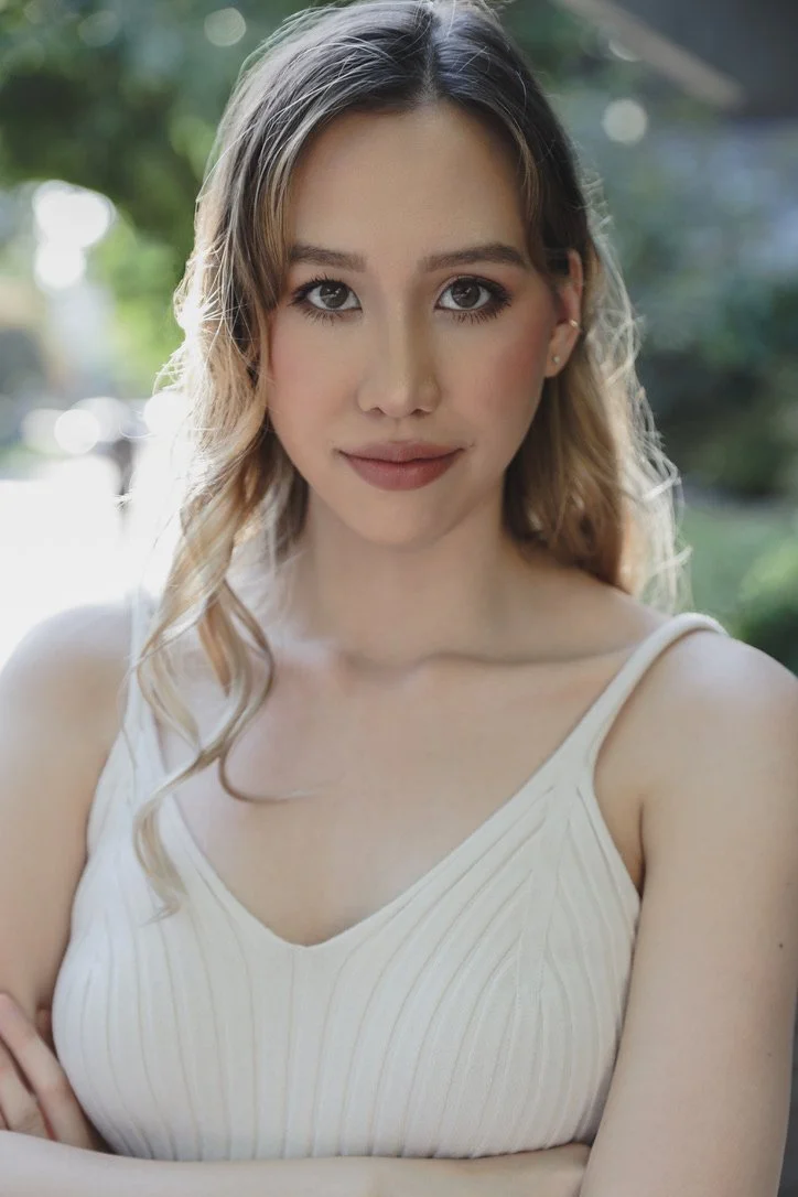 Close-up of a young woman with long wavy hair, wearing a white ribbed tank top, standing outdoors with blurred green trees in the background.