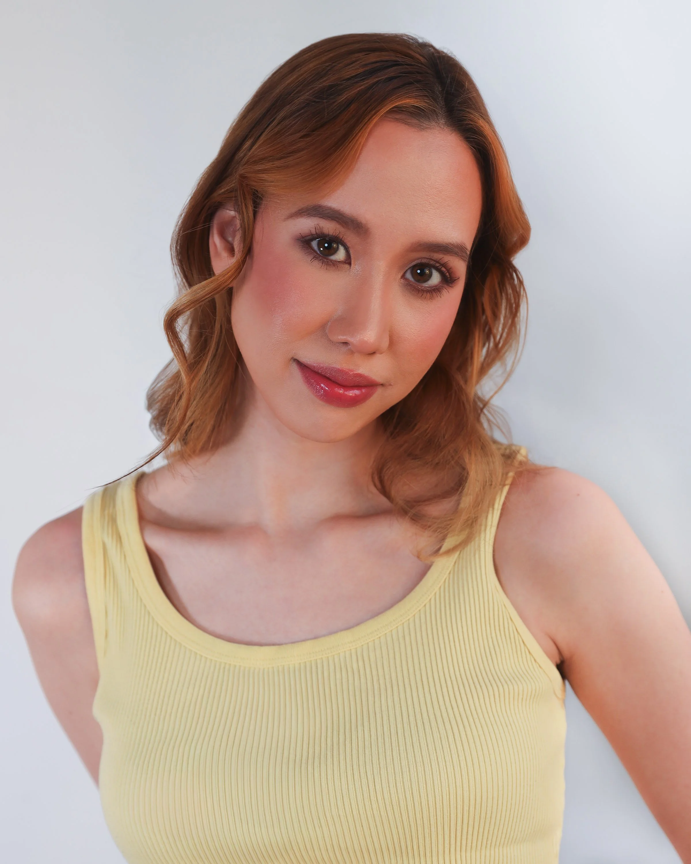 A woman with wavy, shoulder-length reddish-brown hair wearing a sleeveless pale yellow top, standing against a plain white background, smiling softly and looking at the camera.