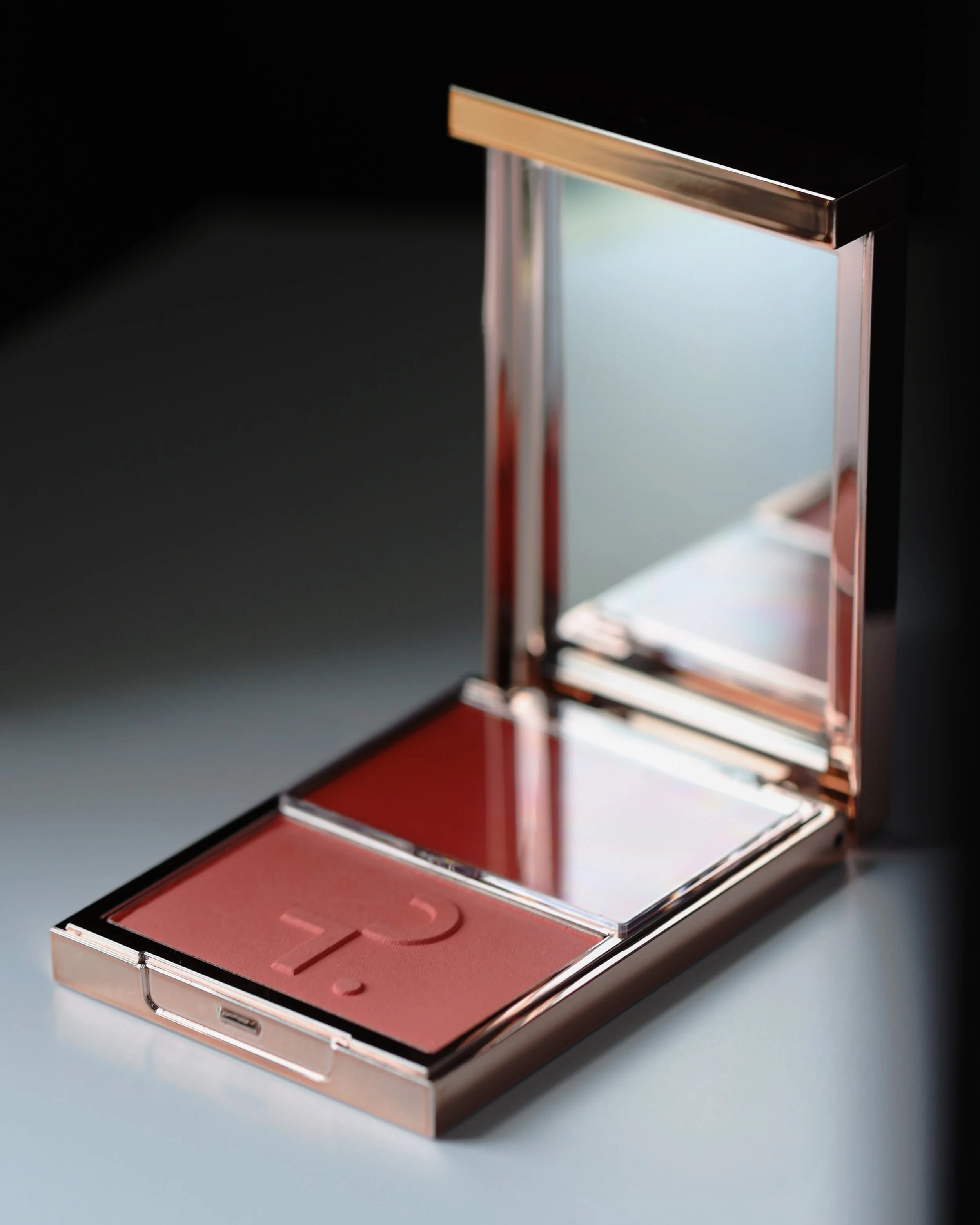Open makeup compact with mirror and pink blush inside, set against a dark background.